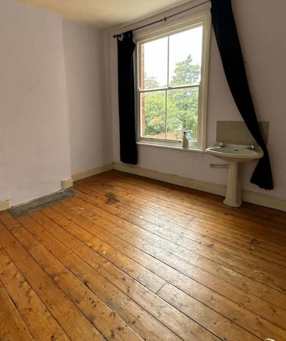 An empty bedroom with bare floorboards, a washbasin in the corner and dark curtains
